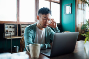 Worried young Asian man with his hand on head, using laptop computer